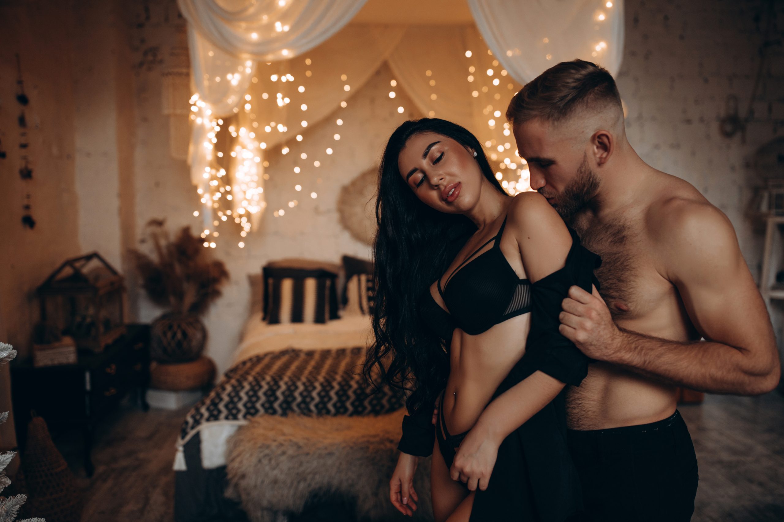 In einem gemütlichen Schlafzimmer steht ein Mann mit freiem Oberkörper und Küsst eine Frau in Unterwäsche auf die Schulter