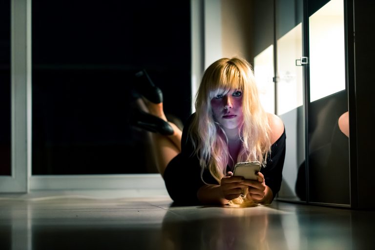 Blonde Frau im sexy Outfit liegt mit Handy auf dem Fußboden und schaut in die Kamera